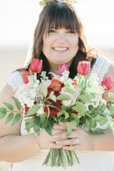 A bride with a big smile holds a bouquet of flowers in front of her body in this close up photo. The bouquet is adorned with red tulips and roses. The woman has long dark hair with bangs covering her eyebrows.