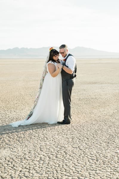 A bride and groom slow dance together on their wedding day. It is a sunny afternoon and they are wearing sunglasses. Their dance floor is the dry and cracked ground of the Dry Lake Bed outside of Las Vegas.