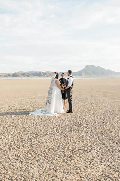 A wide shot of a bride, groom and officiant standing in the middle of the Dry Lake Bed during a wedding ceremony. They are surrounded by nothingness except for the dry cracked ground stretching off into the distance where mountains rise in the background.