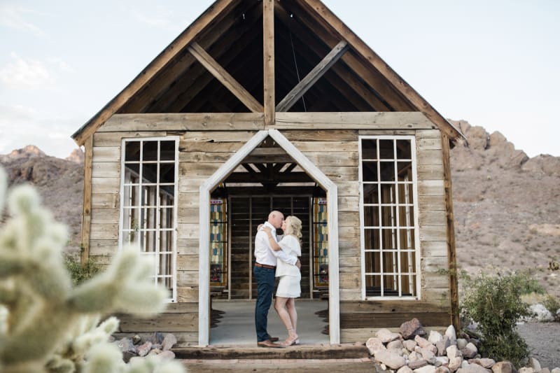 A couple renewing their vows stands in the doorway of a rustic western chapel in Eldorado Canyon near Las Vegas. They are holding each other close and kissing.