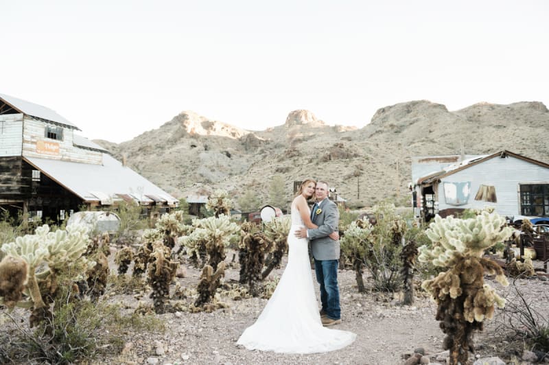 A bride and groom hold each other around the waist while standing in a field of cholla cactus between two old mining buildings in the Eldorado Canyon south of Las Vegas, Nevada.