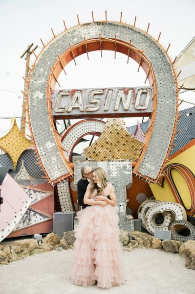 A man and his wife kiss and embrace in front of the old Horseshoe Casino sign which is the standout piece in a pile of large old neon signs at the Neon Museum in Las Vegas.