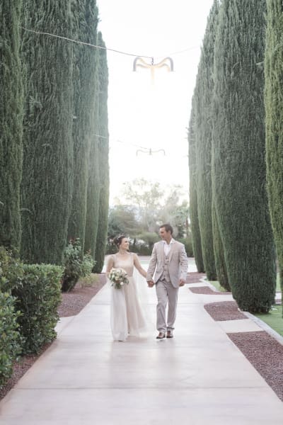 A wide shot of a couple that has just renewed their vows and are walking hand-in-hand through a garden corridor framed by tall Cypress trees. 