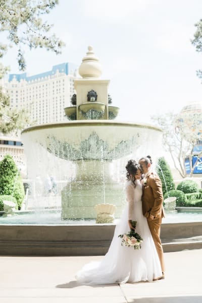 A bride and groom pose for wedding photos on a sunny day in front of an ornate water fountain at the Bellagio Hotel and Casino in Las Vegas. The groom appears to be whispering in the bride's left ear as she holds her flowers down on her right side.