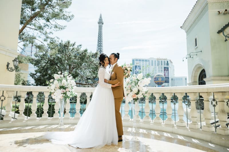 A bride and groom hold each other as the groom nuzzles into the side of his wife's head. They are standing on a balcony overlooking the lake in front of the Bellagio Hotel and Casino in Las Vegas with the Paris Las Vegas's Eiffel Tower in the background.
