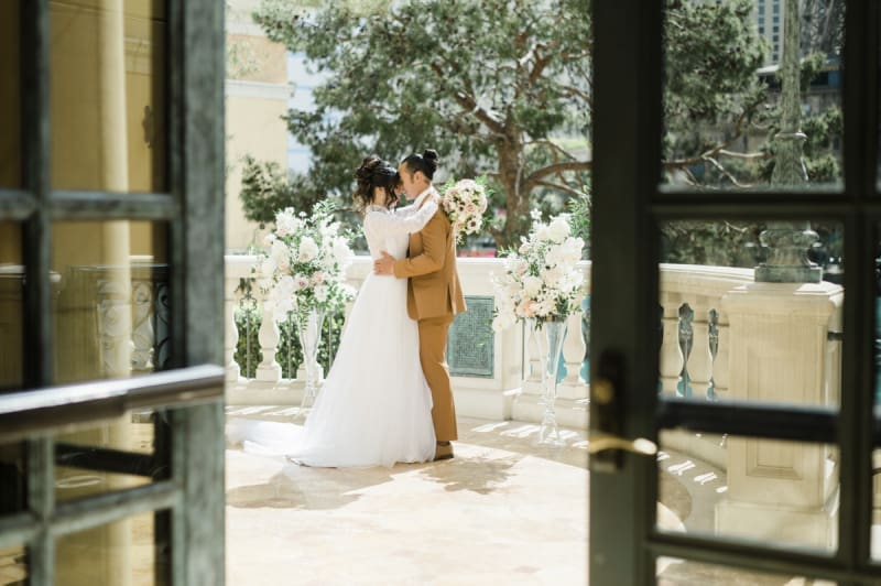 A bride and groom are seen through the open half of a set of French doors. They embrace while standing between two large floral arrangements on a balcony with pine trees in the background.
