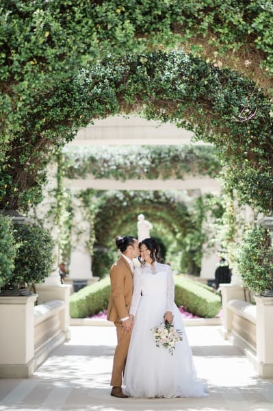 A groom and pride pose underneath a series of formal garden archways on their wedding day. The symmetry of the repeating arches makes for an artistic photograph. The arches are part of the manicured grounds at the Bellagio Hotel and Casino in Las Vegas.