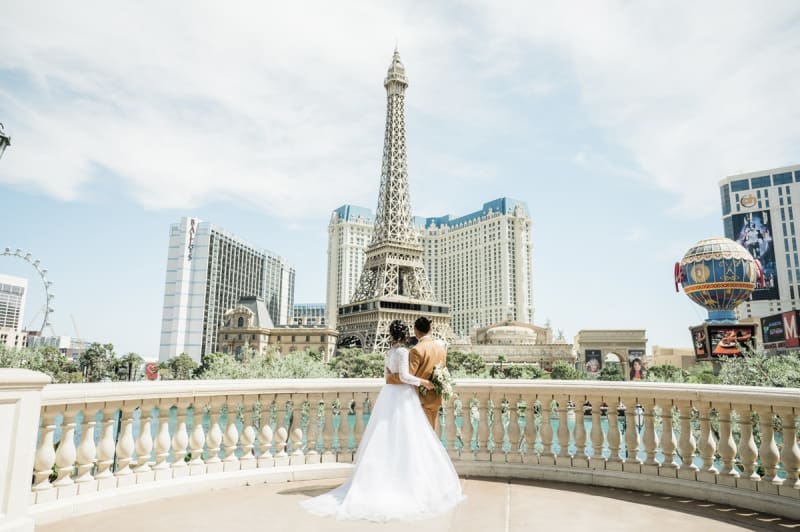 A bride and groom hold each other around the waist while looking out across the Las Vegas Strip. They are standing at a railing which shields the lake at the Bellagio Hotel and Casino in Las Vegas. Parts of The LINQ, the Flamingo, Bally's, Paris, and Planet Hollywood are all visible.