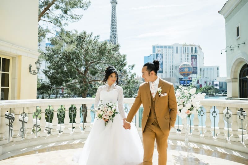 A bride and groom hold hands and look at each other as they step towards the camera. They are on a patio at the Bellagio Hotel and Casino which overlooks the Paris Las Vegas Hotel and Casino and Planet Hollywood Las Vegas Resort and Casino.