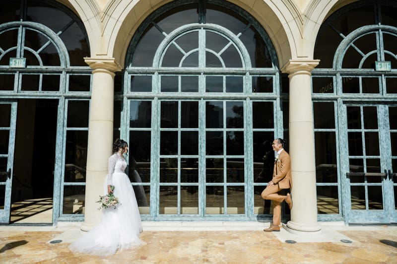 A bride and groom lean against Tuscan columns that frame a row of arched glass windows. They look at each other across the space that separates the two columns. They are posing for photos on their wedding day at the Bellagio Hotel and Casino in Las Vegas.