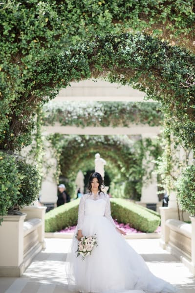 A wide shot of a bride in a white wedding dress standing underneath a series of formal garden archways. The arches are covered in climbing plants which obscure the structure holding them up.