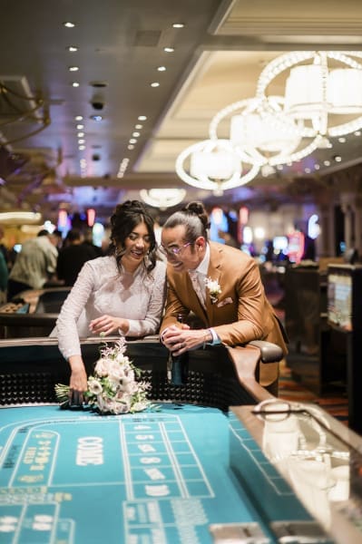 A bride and groom share a laugh at an empty craps table inside the Bellagio Hotel and Casino in Las Vegas. Her wedding dress has long sleeves, and he wears a brown suit. Modern spherical light fixtures hang above them in the out of focus background.