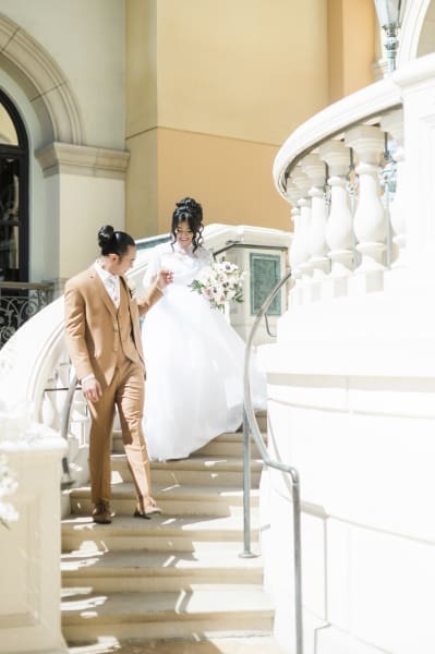 A groom leads his bride down a white, curved, Italianate staircase. He holds her hand to assist her as she smiles and watches her step. He wears a brown suit. She wears a long white dress and carries a floral bouquet.