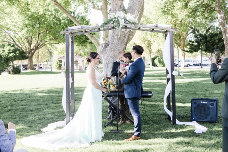 A bride and groom stand underneath an arbor set up under a big tree in a park. The groom holds a small boy.