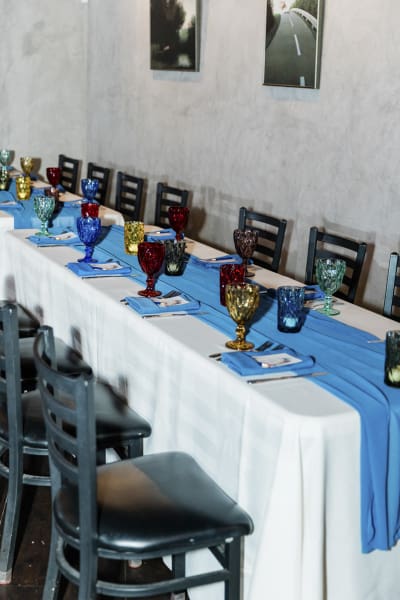 A long communal dining table is set up for a reception. There is a blue runner atop a white tablecloth.