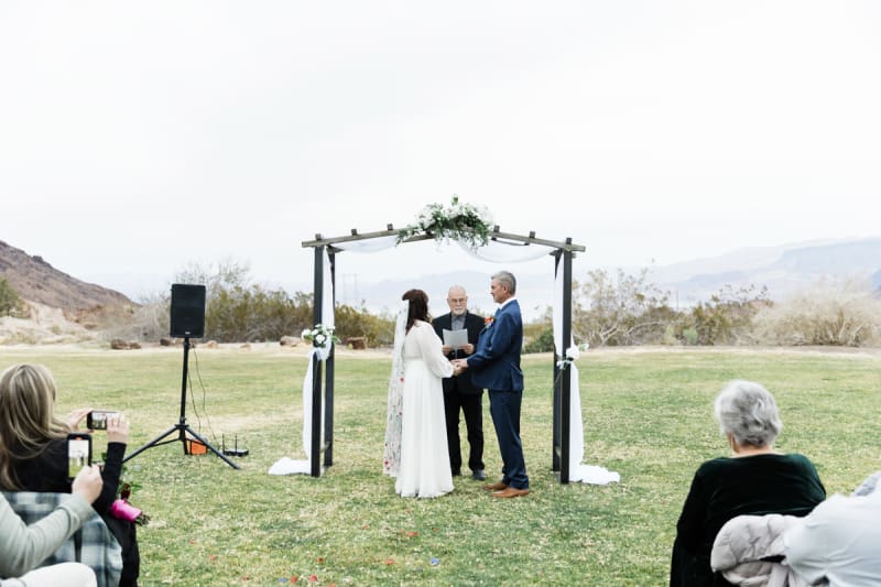 A wedding is held on a green lawn at a park in Boulder City with mountains in the background.