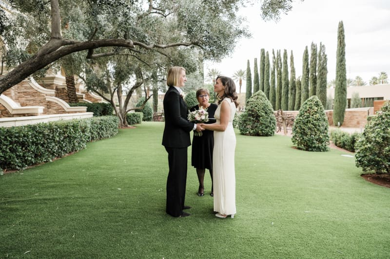 Two women hold hands and smile at each other as they are married by an officiant in a garden at Green Valley Ranch.