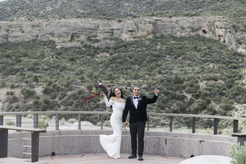 A bride and groom raise their arms in celebration of their marriage at Lee Canyon outside of Las Vegas as they stand in front of a hillside dotted with a large rock outcropping.