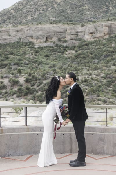 A bride and groom kiss and hold hands as they stand and face each other with a mountain hillside in the background.
