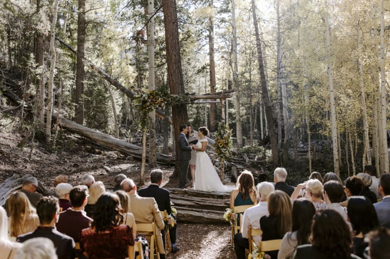 Wide shot of an outdoor wedding taking place in the mountains at Lee Canyon near Las Vegas. The wedding guests are seated on wooden folding chairs as they watch the bride and groom at a natural wooden arbor covered with wedding flowers.