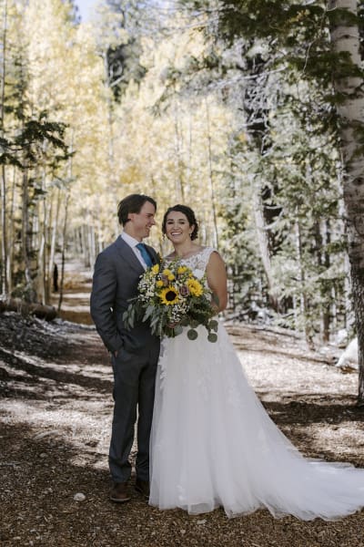 A smiling bride and groom pose for the camera while standing on a dirt road carved through the pine trees and aspens in the mountains on their wedding day.