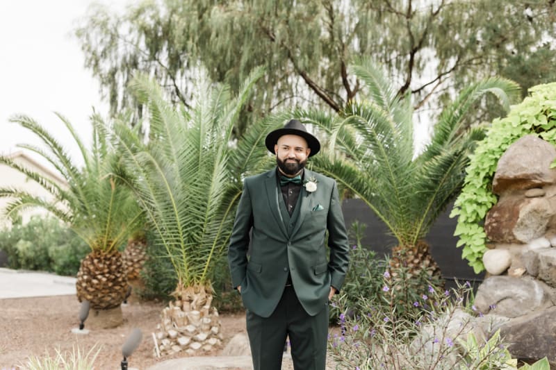 A groom in a dark green suit and a black hat poses for a wedding portrait in the garden at Lotus House in Las Vegas.