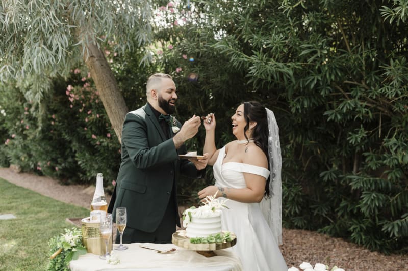 A groom and bride laugh as they hold wedding cake forks towards each other. They are in a garden.