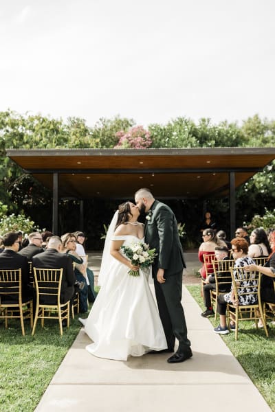 A bride and groom kiss. They are standing at the rear of all their seated wedding guests in a garden.