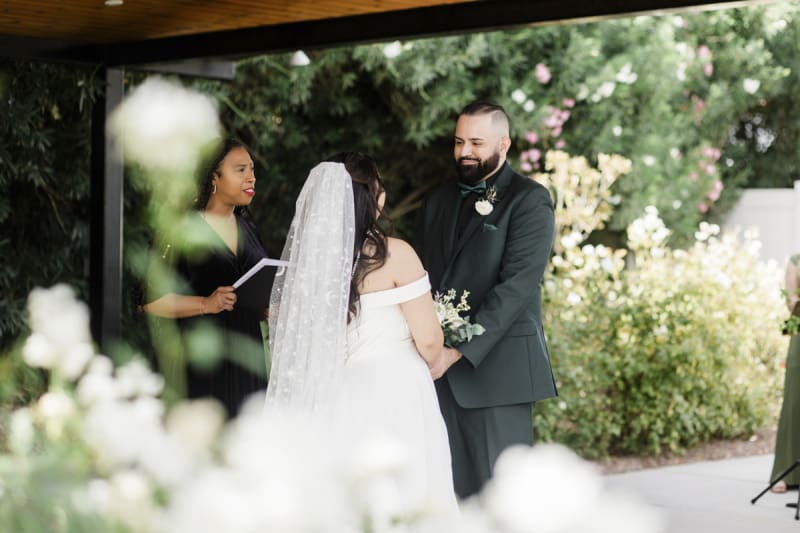During a wedding ceremony, a groom looks at his bride who has her back to the camera. There is a rose garden in the background.