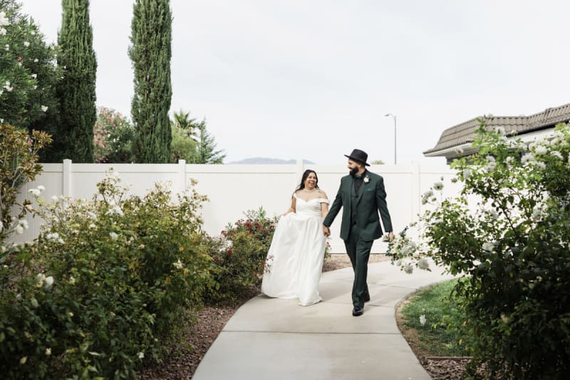 A bride and groom happily walk down a curved sidewalk that winds between white rose bushes.
