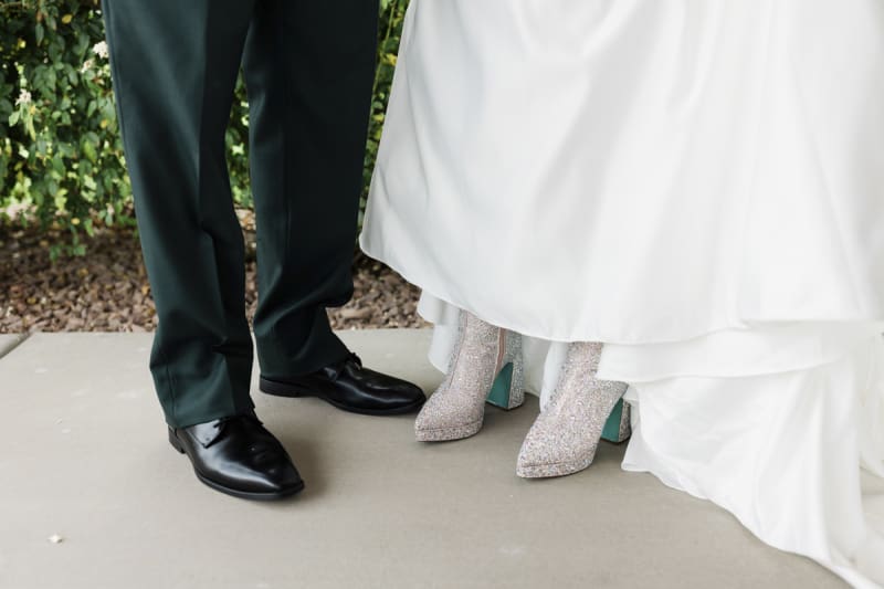 A closeup of the feet of a groom in black pants and black shoes, and a bride in a long floor length dress with sparkly silver boots.