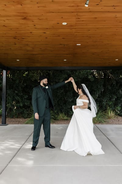 A groom twirls his bride underneath a pergola with a warm-colored wooden roof.
