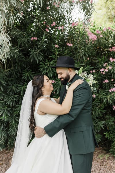 A bride and groom hold each other and smile as they stand in front of a green and pink hedge.