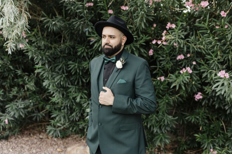 A medium shot of a man with a beard, a green suit and a black fedora standing against a hedge of pink Oleander bushes.