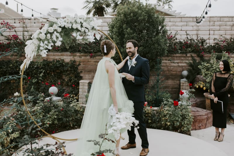 A bride puts her left hand on her groom's shoulder as he reads his wedding vows to her.  The wedding officiant stands to the side and watches as the couple stands in front of a round wedding arbor in the middle of a rose garden.