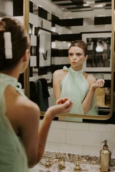 A bride in a light mint colored wedding dress puckers her lips in front of a mirror. The mirror and other bathroom fixtures are a bright bronze color while the wall art in the reflection is black and white.