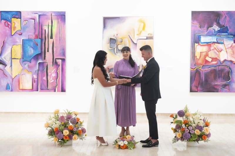 A bride places a wedding ring on her groom's finger during their wedding ceremony inside an art museum. as the officaint looks on.