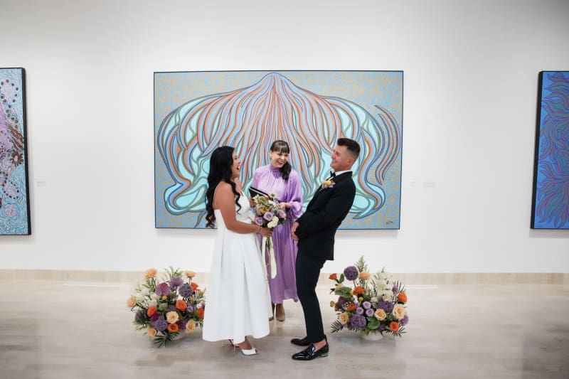 A bride, groom and officiant laugh during a wedding ceremony in front of a large abstract painting in an art gallery.