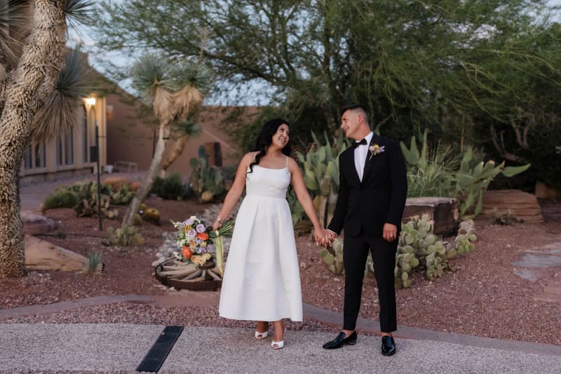 A bridge and groom hold hands and plyfully look at each other at dusk in a cactus garden.