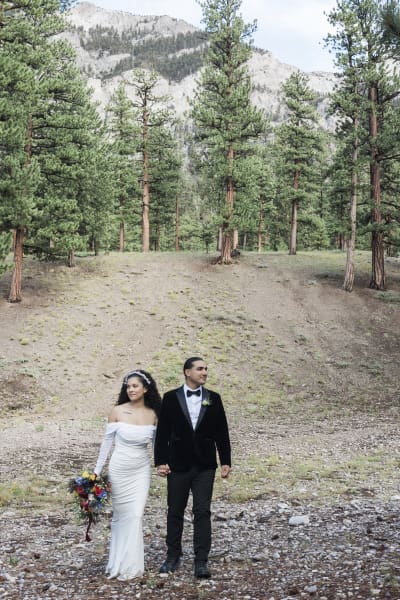 A bride and groom walk down a gentle mountain slope holding hands. The bride holds her multi-colored floral bouquet in her outside hand.