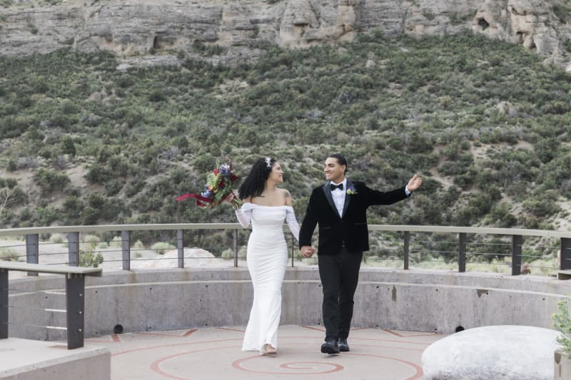 A bride and groom walk towards the camera and look at each other gleefully  during their wedding in the Spring Mountains outside of Las Vegas. The hillside behind them is dotted with a large rock outcropping.