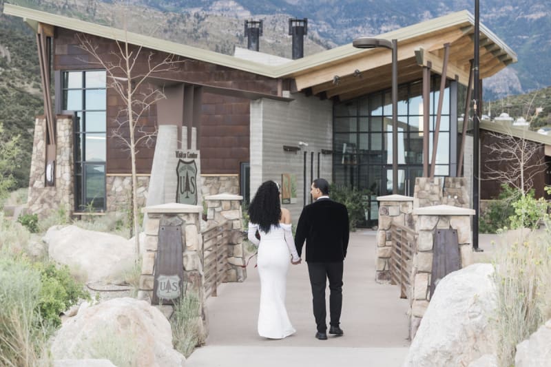A bride and groom walk hand-in-hand away from the camera and towards the visitor center building at the Spring Mountains Visitor Gateway. They pass through a bridgeway built of native stone. 