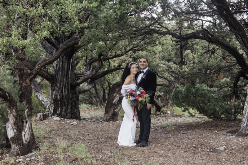 A bride and groom smile and pose for wedding photos in front of craggy old pine trees. The bride holds a bouquet of flowers with a dark red ribbon dangling from it.