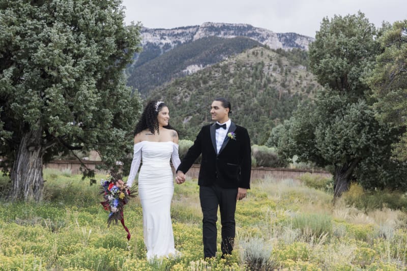 A bride and groom hold hands and turn their heads to look at each other as they stand in a meadow of wildflowers between pine trees with tree covered mountains in the background.