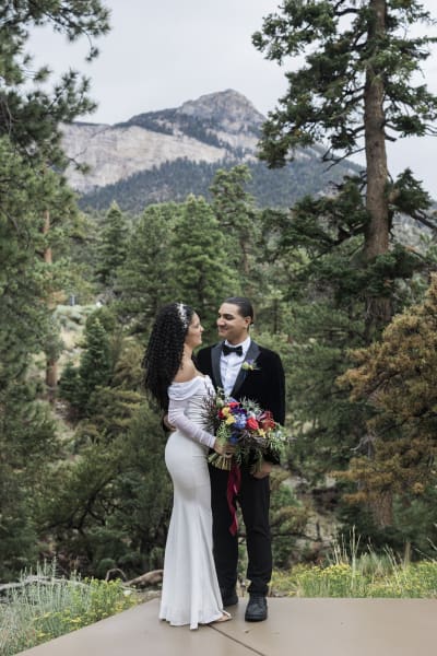 A bride and groom stand closely together at the edge of a paved patio that overlooks a forest of tall pine trees and a mountain peak.