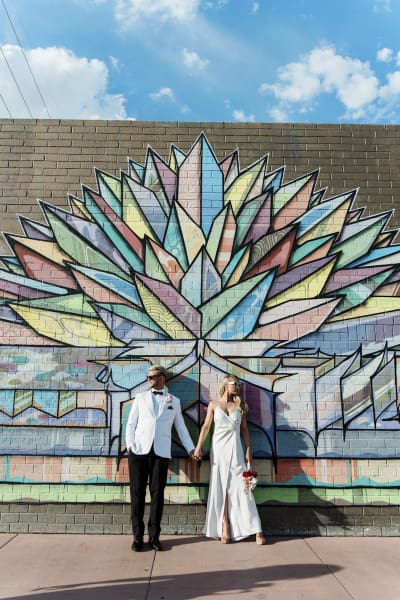 A bride and groom posing in front of a brick mural. They mural is painted with a colorful geometric crystal 