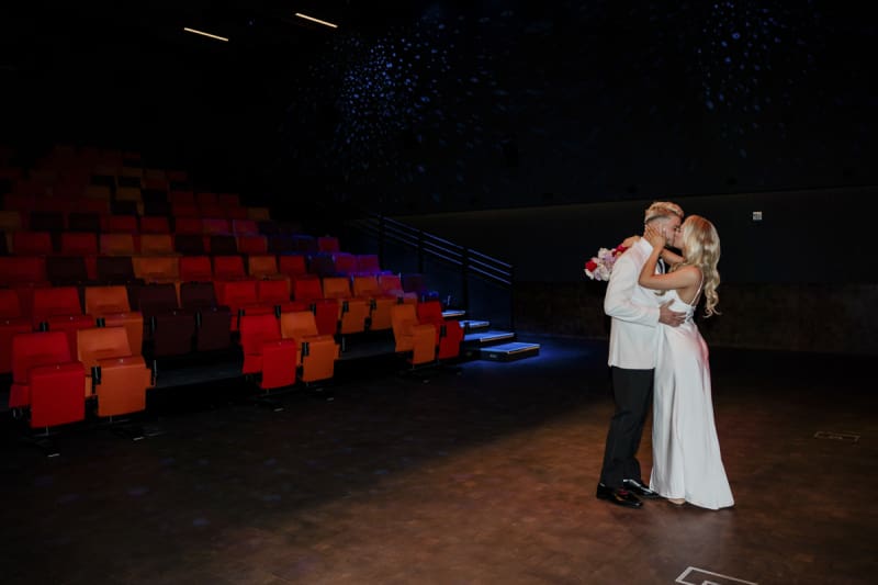 A bride and groom kiss as they dance in the empty Beverly Theater main room. The red seats are illuminated by blue light in this moody looking photo.