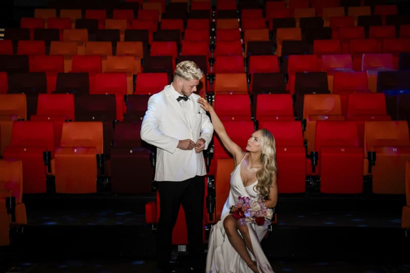 A standing groom buttons his white tuxedo jacket as a seated bride reaches up to adjust his collar. They are getting married at the arthouse theater in Downtown Las Vegas.