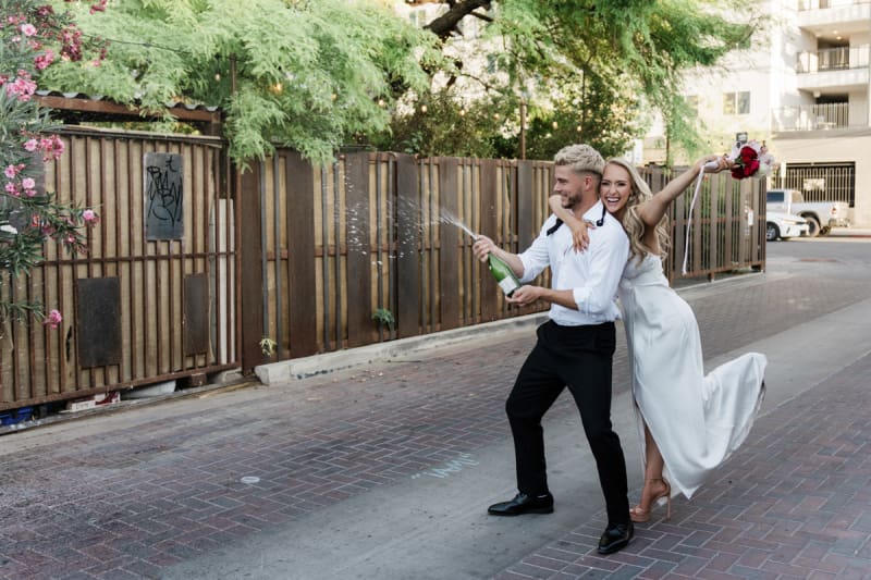 A bride grabs hold of a groom while the groom sprays Champagne from a bottle. They are posing for pictures in a fence-lined alley in Downtown Las Vegas.
