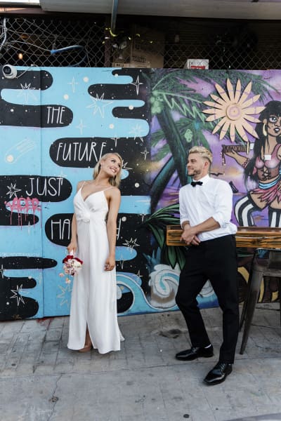 A groom leans against a high-top table as he looks at his bride. The bride turns back dramatically to look at the groom. They are in front of a colorful mural in Downtown Las Vegas.